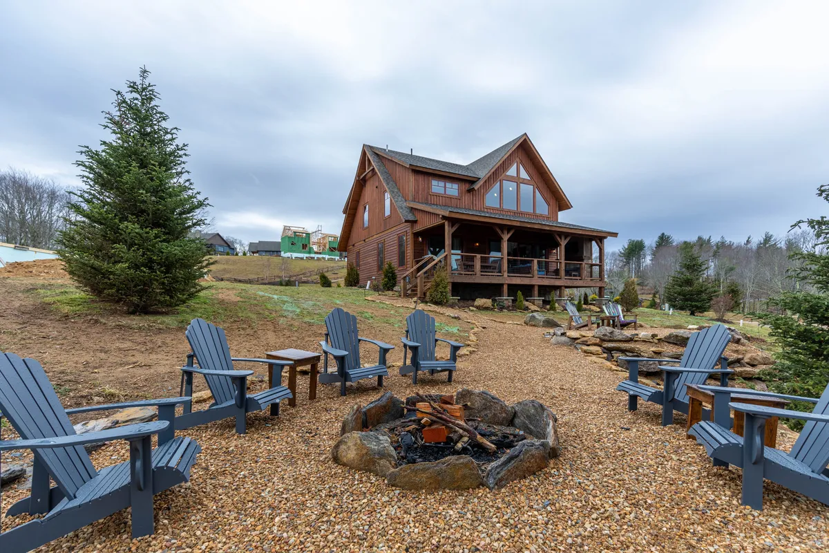 The hero image features a spacious log cabin nestled among tall trees, with a welcoming fire pit encircled by Adirondack chairs, all set against the backdrop of lush mountain scenery. The outdoor setup invites guests to gather, relax, and enjoy the peaceful natural surroundings as the sun sets over the mountains.