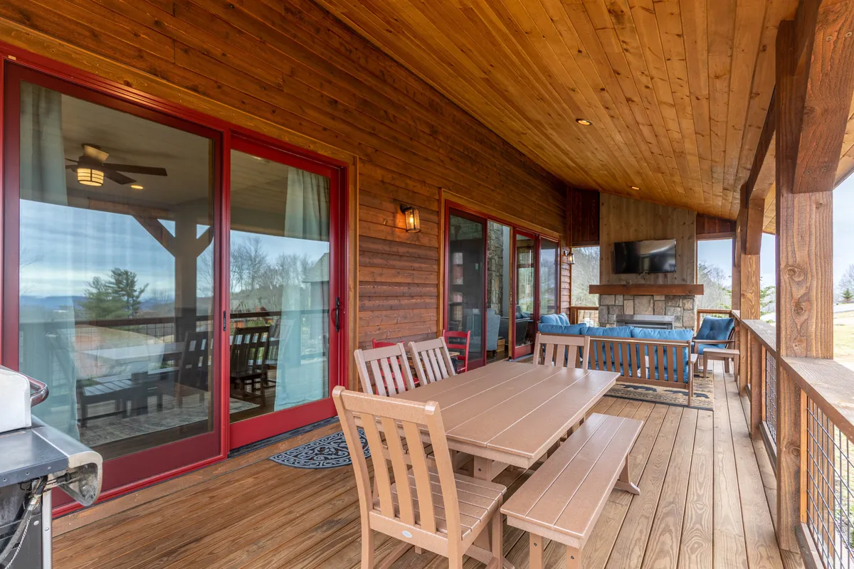 An inviting exterior dining area features a large wooden table, red-framed sliding doors, a cozy outdoor fireplace, and plush seating. The space is set against a backdrop of natural mountain views, with a grill ready for al fresco meals and gatherings, blending resort comfort with the outdoors.