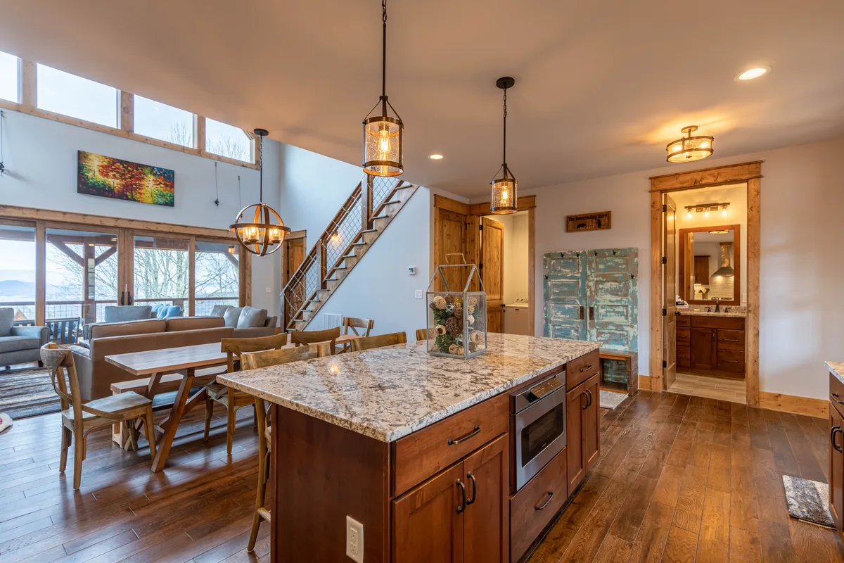 An open-concept kitchen and dining area with a large granite island, a wooden staircase, and a sunlit dining table positioned by expansive windows. The inviting feel encourages both cooking and communal dining or game nights.