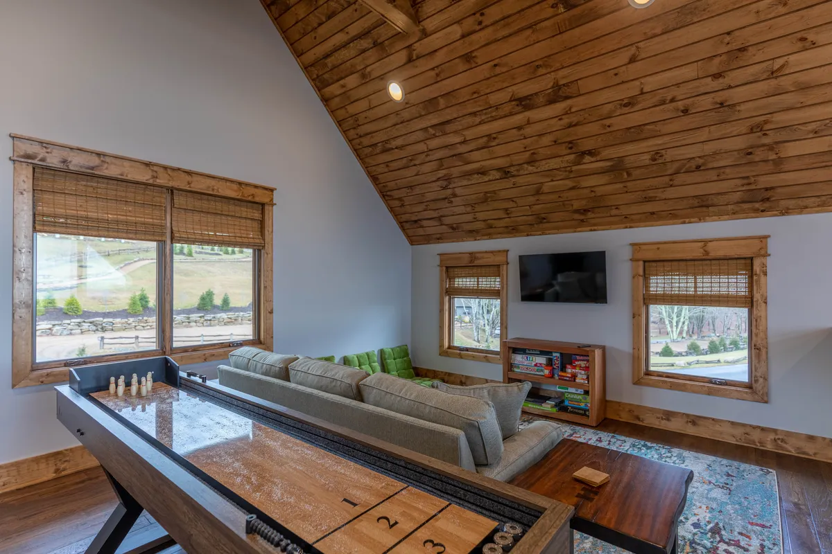 A cozy living room corner featuring a shuffleboard table, comfortable sofa, bookshelves, windows letting in natural light, and a television mounted on the wall. The setting feels playful yet relaxing—ideal for games, movie nights, and shared downtime.