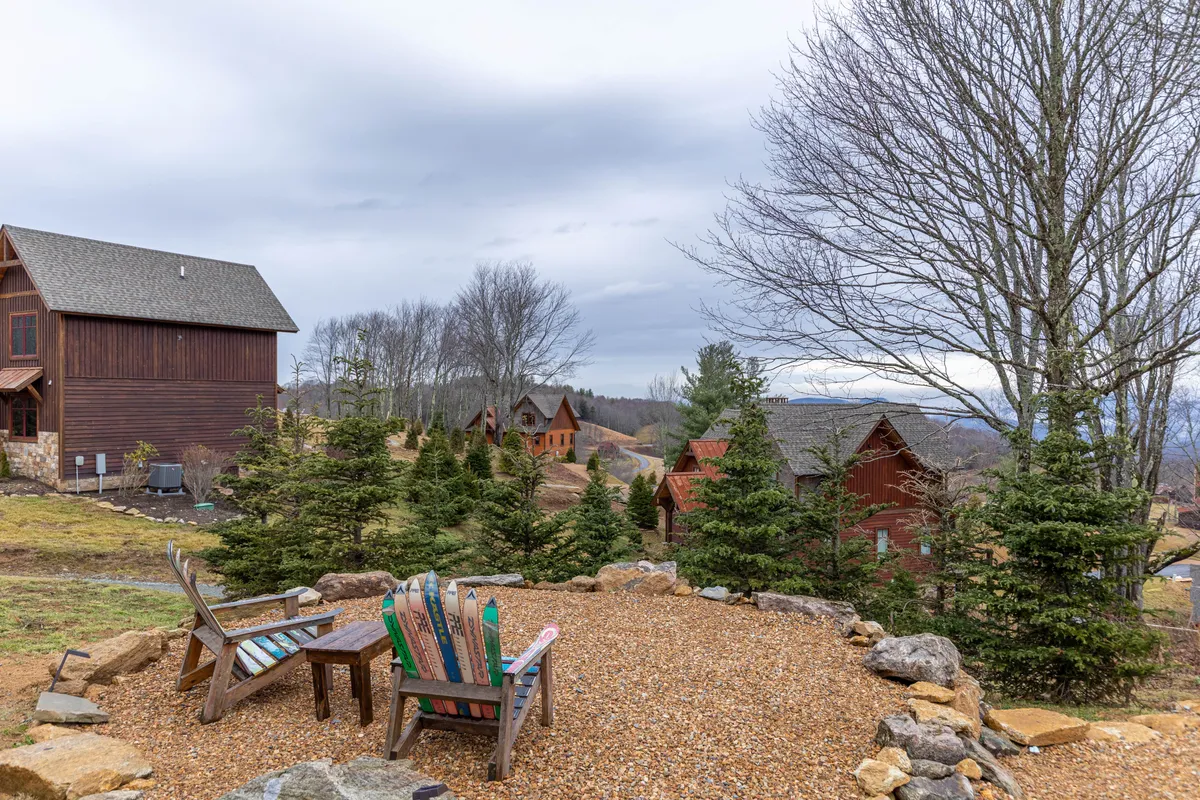 An inviting outdoor seating area featuring colorful Adirondack chairs arranged around a natural rock garden, surrounded by tall pine trees and with distant cabins visible in the background. The scene is tranquil yet full of potential for group gatherings and relaxation in nature.
