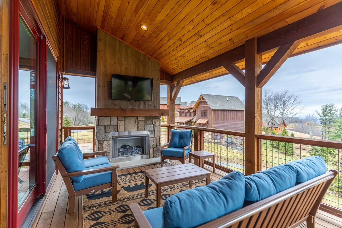 This image showcases a cozy exterior covered deck with a fireplace, plush seating, a wooden ceiling, and a scenic landscape view beyond the railing. The atmosphere is inviting, warm, and ideal for year-round relaxation or entertaining guests.