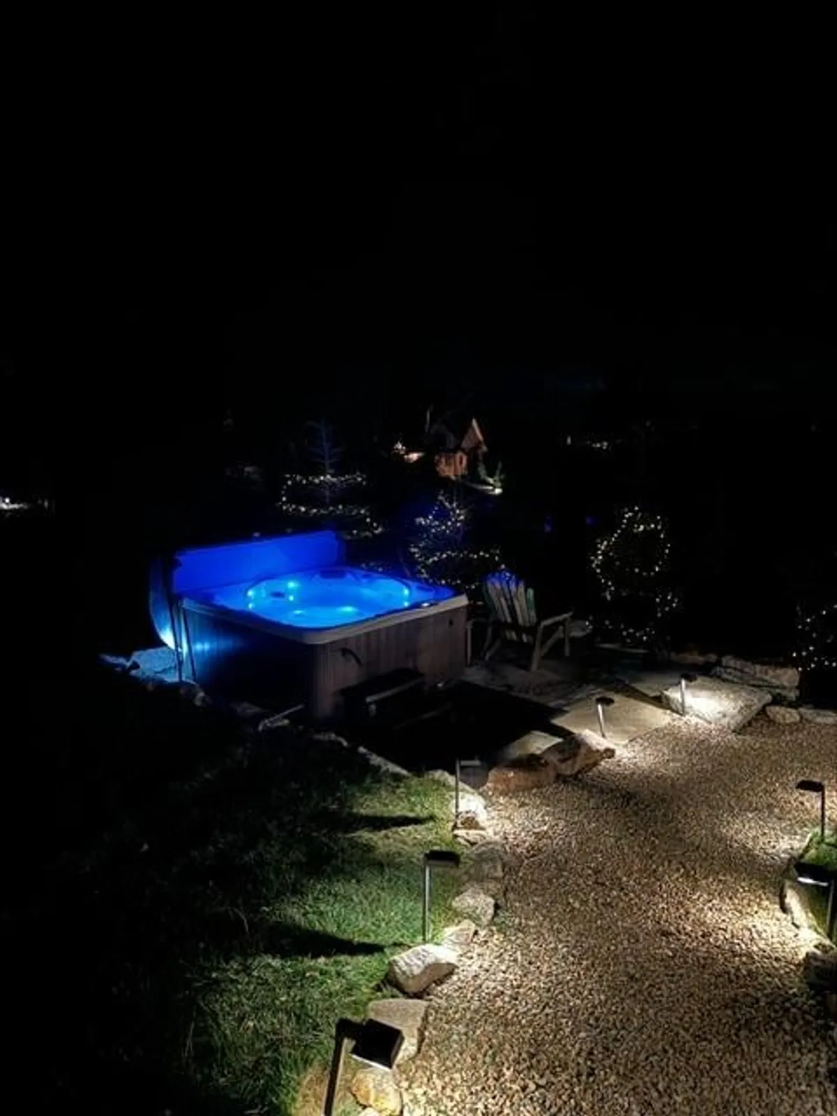 The image features a hot tub illuminated with blue lighting, surrounded by adirondack chairs and trees adorned with twinkling lights. The setting suggests a serene, magical atmosphere ideal for relaxation or intimate gatherings under the stars.