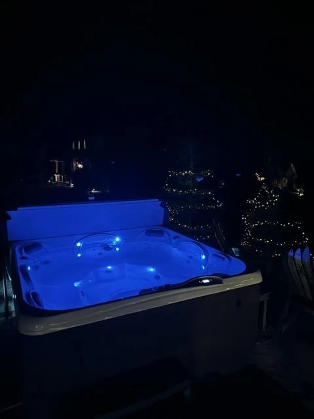 An exterior shot of a hot tub lit by blue lights, surrounded by twinkling string lights, suggesting a festive and inviting place for families or groups to relax outdoors, especially in the evening.