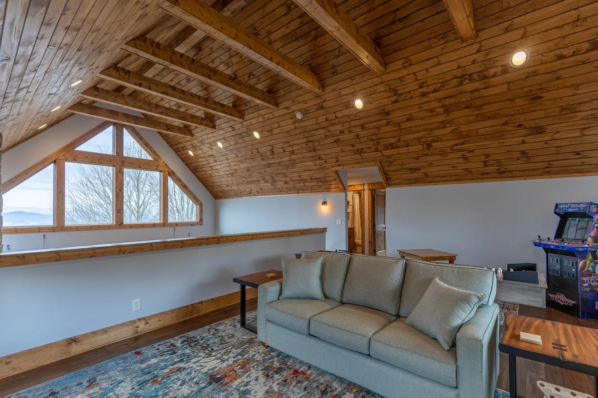 A spacious area with large windows framing the mountain landscape, exposed wooden beams, and a comfortable couch. There's also a game arcade machine, suggesting opportunities for group fun and entertainment in a setting with beautiful outside views.