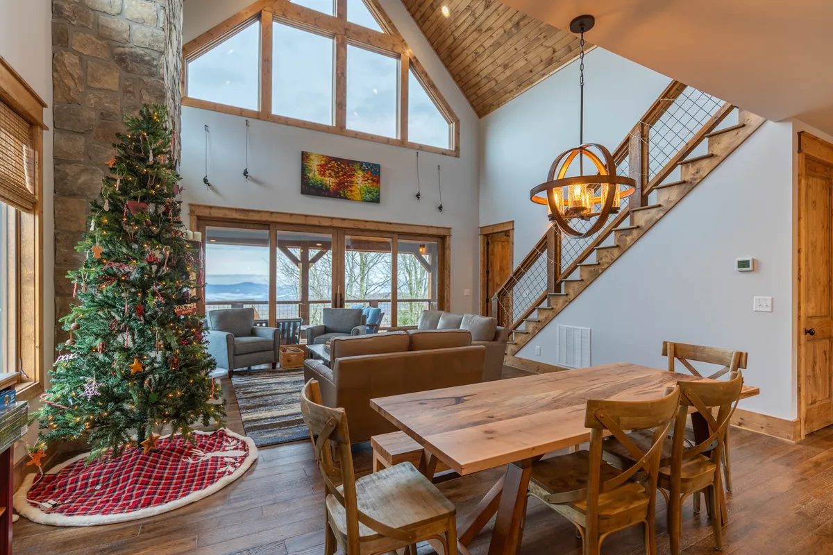 A bright and inviting interior space featuring large windows that offer a glimpse of the surrounding mountain landscape. The room is arranged for comfort with a cozy seating area, a wooden staircase, a festive Christmas tree, and a dining table, suggesting ample space for relaxation and togetherness. The abundance of natural light and outdoor views hint at the connection to nature just beyond the home.