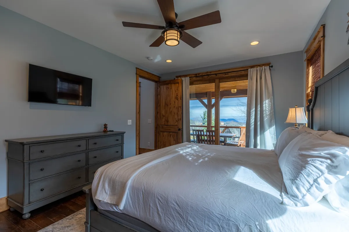 A tranquil king bedroom with a plush bed dressed in crisp white linens, a wooden dresser, and a large window that frames a scenic mountain view. The soft lighting and serene atmosphere invite relaxation, while the window brings the beauty of nature indoors.