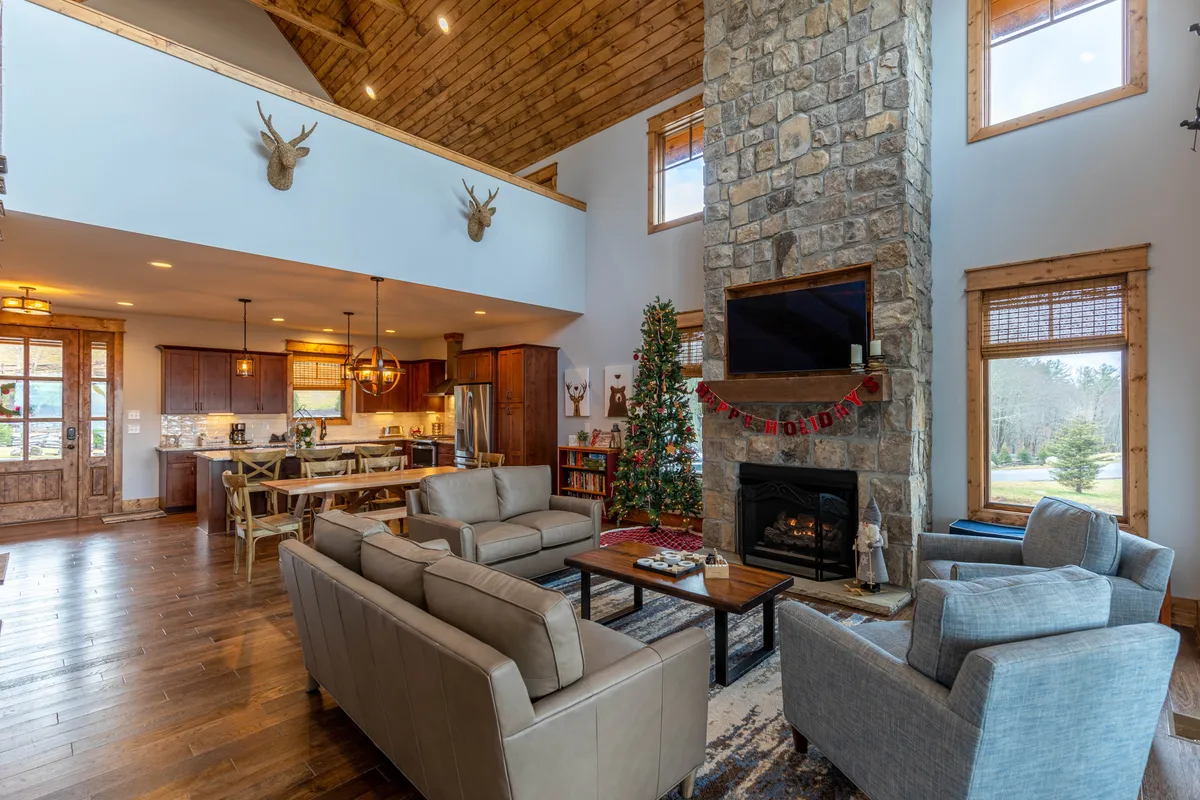 A cozy living area with a stone fireplace as the focal point, surrounded by comfortable sofa seating and large windows that reveal natural light. A decorated Christmas tree hints at festive celebrations and the changing seasons outside.