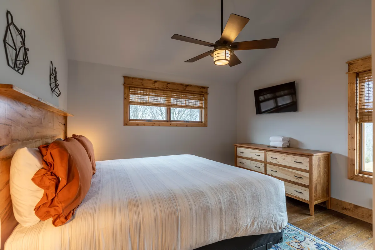 A restful King bedroom with a large, comfortable bed, ceiling fan, windows letting in gentle daylight, a dresser, and a television. The setup is inviting for both a good night's sleep and leisurely afternoons watching TV or reading.