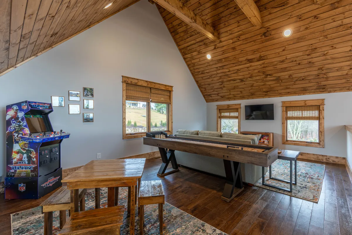 A spacious recreation room featuring a classic arcade game machine, a large wooden table for games or dining, a comfortable couch for lounging, and walls adorned with photos. The atmosphere is playful and social—perfect for indoor fun regardless of weather.